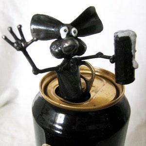 May include: A black metal sculpture of a cartoon dog with large ears and googly eyes, holding a black metal can. The dog is sitting on top of a black can with a gold top, which has the word "Guinness" printed on it.