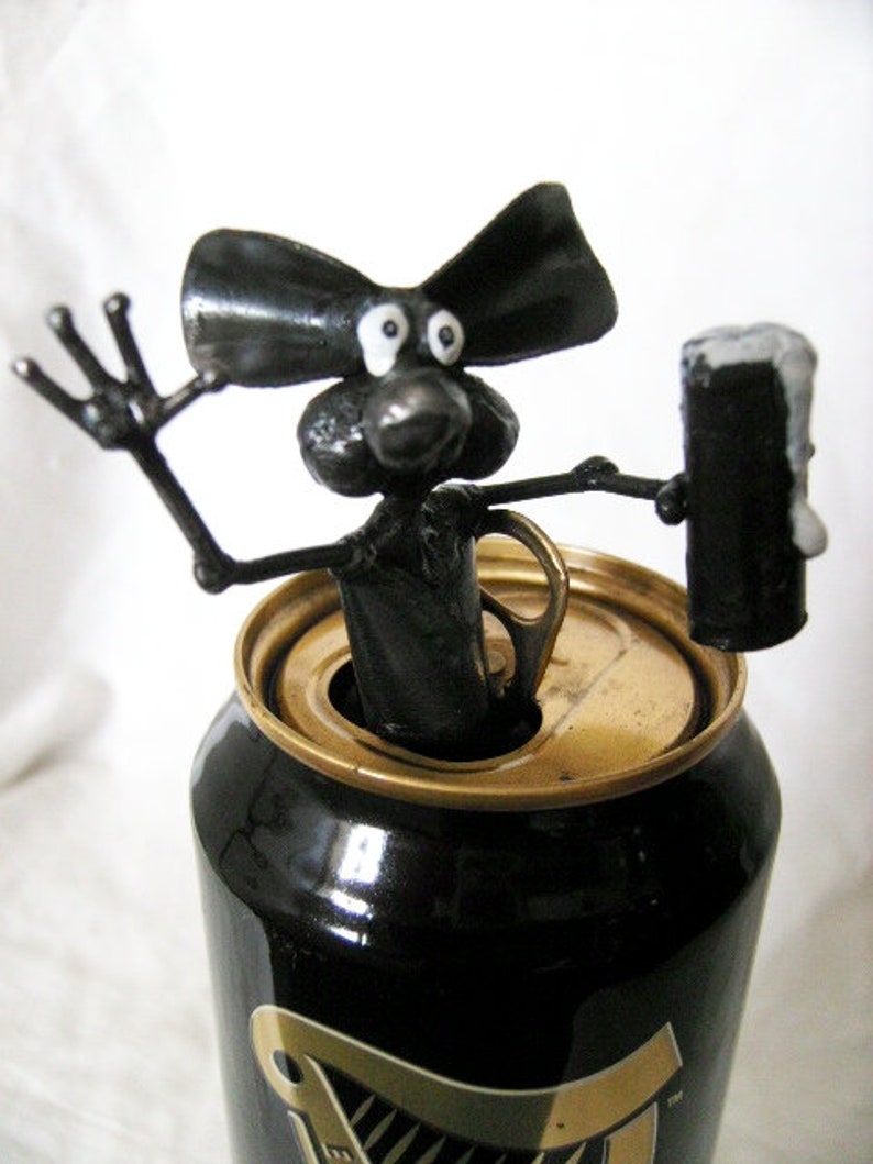 May include: A black metal sculpture of a cartoon dog with large ears and googly eyes, holding a black metal can. The dog is sitting on top of a black can with a gold top, which has the word "Guinness" printed on it.