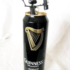 May include: A black and gold Guinness can with a black metal sculpture of a dog holding a can of Guinness on top. The can says "Guinness Draught" and "Brewed in Dublin".