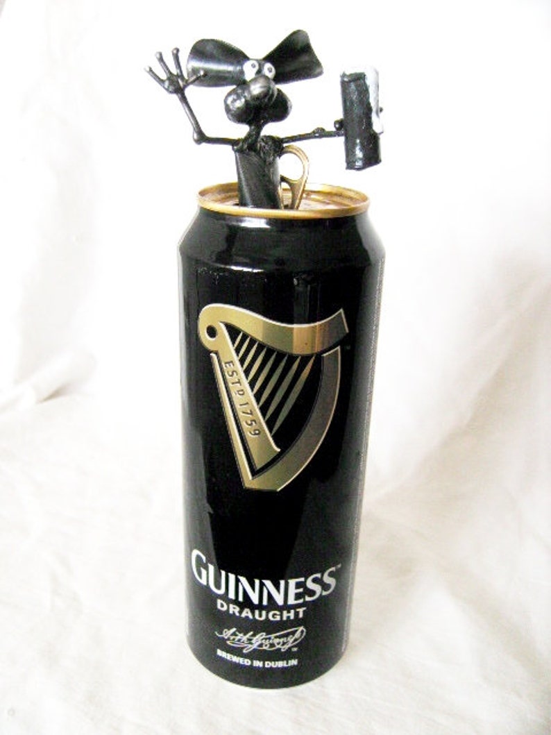 May include: A black and gold Guinness can with a black metal sculpture of a dog holding a can of Guinness on top. The can says "Guinness Draught" and "Brewed in Dublin".