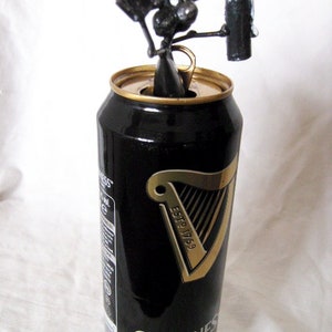 May include: A black metal sculpture of a person holding a can of Guinness Draught beer. The sculpture is sitting on top of the can, which is closed and has a gold pull tab.