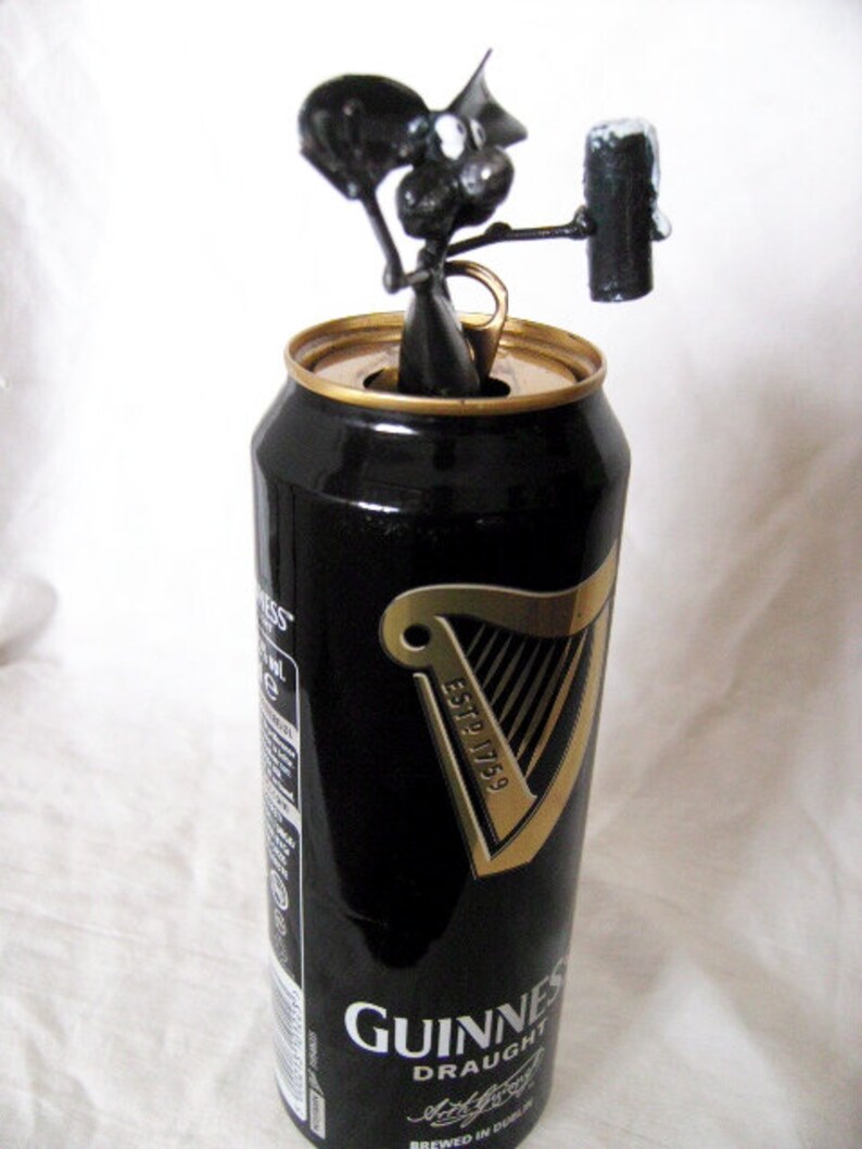 May include: A black metal sculpture of a person holding a can of Guinness Draught beer. The sculpture is sitting on top of the can, which is closed and has a gold pull tab.