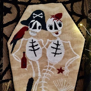 May include: Cross-stitch embroidery of two skeletons in a coffin. The skeleton on the left is wearing a pirate hat and holding a bottle. The skeleton on the right is wearing a red hat and has a fishtail. The background is a beige fabric with a spiderweb pattern.