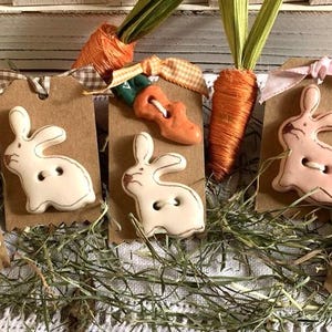 Puede incluir: Seis botones de cerámica con forma de conejo con detalles de zanahoria. Los botones están unidos a etiquetas de papel marrón con cuerda y cinta. Los botones son blancos, rosas y crema.