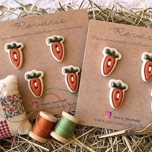 May include: Two cards of handmade Italian ceramic carrot-shaped embellishments. Each carrot is orange with a green top and white outline. A patchwork pin cushion, spools of thread, and gold scissors are also visible.