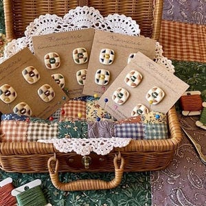 May include: A wicker basket holds several cards of handmade Italian ceramic buttons. Each card displays a set of four square buttons with colorful geometric designs. Spools of thread and scissors are also visible.
