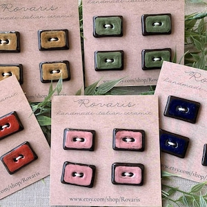 Op de afbeelding: Handgemaakte Italiaanse keramische knopen in diverse kleuren, waaronder rood, roze, groen, blauw en beige. Elke rechthoekige knoop heeft een donkerbruine rand en een wit draadgat. De kaarten lezen "Rovaris handmade italian ceramic."