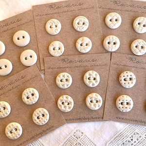 Könnte beinhalten: Cremefarbene Keramikknöpfe auf braunen Karten. Die Knöpfe haben verschiedene geprägte Designs und zwei oder vier Löcher zum Annähen. Die Karten sind mit "Rovaris handmade italian ceramic" beschriftet und enthalten eine Shop-URL.