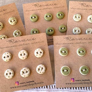 Op de afbeelding: Verschillende kaarten met handgemaakte Italiaanse keramische knopen in diverse ontwerpen. Sommige knopen zijn olijfgroen met witte accenten, andere hebben geruite of gestippelde patronen. De kaarten zijn gemarkeerd met "Rovaris handmade italian ceramic".