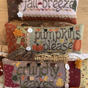 Op de afbeelding: Drie decoratieve kussens met herfst-borduurwerk. Het bovenste kussen is roze met de tekst "fall breeze". Het middelste kussen is groen met de tekst "pumpkins please". Het onderste kussen is grijs met de tekst "crunchy leaves".