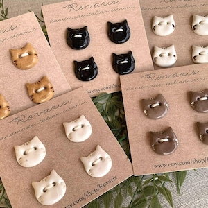 May include: A selection of ceramic cat-shaped buttons in black, white, tan, and brown, presented on tan cards. Each button features two sewing holes. The cards are printed with "Rovaris handmade italian ceramic" and a website address.