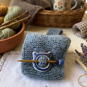 Puede incluir: Un cuadrado tejido azul con un alfiler en forma de gato. El alfiler es blanco y azul. Un cuenco de terracota contiene lana en tonos marrones y verdes. Las agujas de tejer se colocan en el cuenco. Una cesta, tela y flores secas están en el fondo.