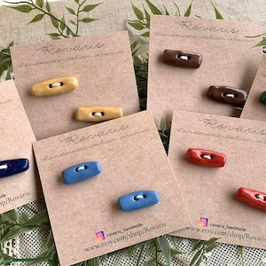 Pode incluir: Uma coleção de botões de cerâmica italiana feitos à mão em várias cores, incluindo azul, amarelo, castanho, vermelho e verde. Cada botão é exibido em um pequeno cartão com o logotipo Rovaris e o link da loja Etsy.