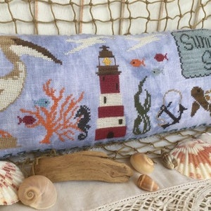 May include: A blue and white embroidered cushion with a nautical theme. The cushion features a pelican, a lighthouse, a sea turtle, coral, a seahorse, seaweed, an anchor, and the words "Summer Set".