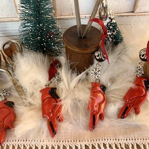 May include: Four red cardinal bird ornaments with silver snowflake charms and black beads hanging from white feathers. The ornaments are attached to red ribbon and are ready to be hung on a Christmas tree.