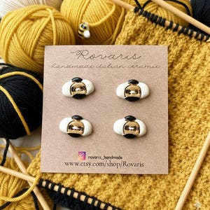 Könnte beinhalten: Vier handgefertigte italienische Keramikknöpfe in Bienenform. Die Knöpfe sind weiß mit goldenen und schwarzen Akzenten. Die Knöpfe werden auf einer braunen Karte mit dem Text "Rovaris handmade italian ceramic" präsentiert. Im Hintergrund sind gelbe und schwarze Wolle und Stricknadeln.