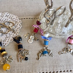 Op de afbeelding: Een set van zes kleurrijke garenrolcharms met verschillende versieringen, waaronder kralen, linten en metalen charms. De charms zijn bevestigd aan zilveren kettingen en worden getoond op een glazen standaard met een verzameling scharen.
