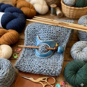 May include: A collection of knitting supplies on a wooden surface. Includes skeins of yarn in various colors, knitting needles, a blue bird-shaped shawl pin, a knitted scarf, gold scissors, and stitch markers. The yarn colors range from blue and brown to green and cream.