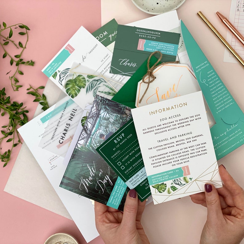K&ouml;nnte beinhalten: Ein Satz Hochzeitseinladungen mit tropischem Thema. Die Einladungen sind gr&uuml;n, wei&szlig; und gold mit einem botanischen Design. Der Text auf den Einladungen lautet "Made our Day" und "Together with their families, Charis Neil".