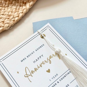 May include: White card with a gold inscription that reads "Happy Anniversary" and a small gold heart. The card has a white tassel attached to it with a gold grommet. The card also has the text "Mrs Becky Ashbee" and "1 Year" printed on it.