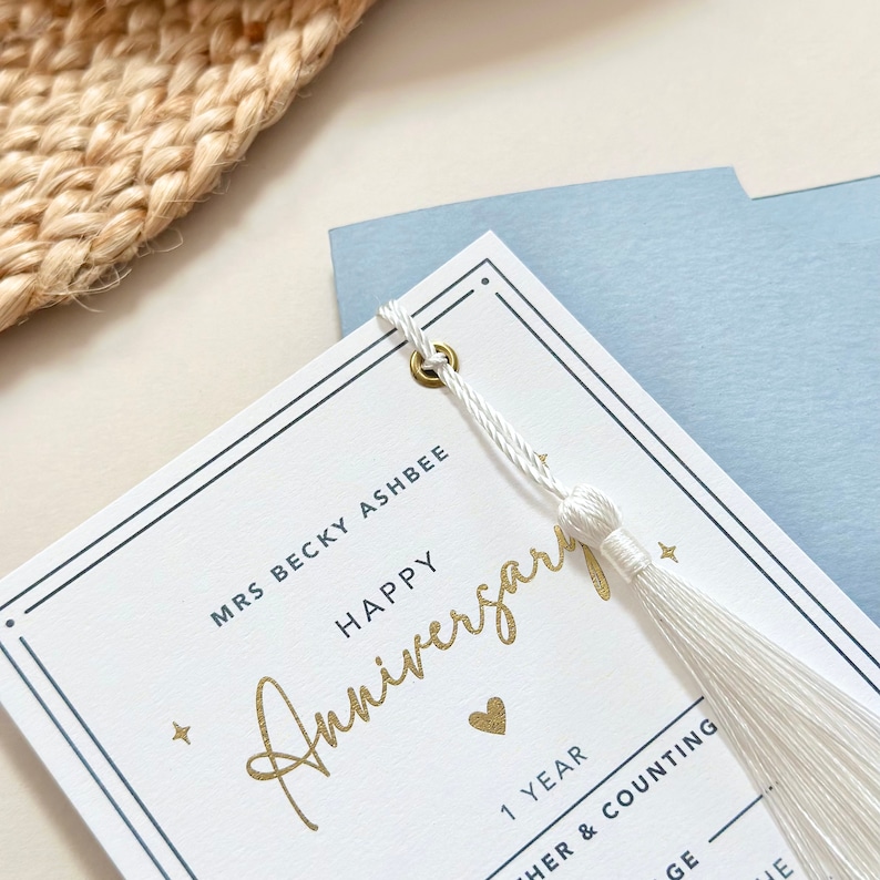 May include: White card with a gold inscription that reads "Happy Anniversary" and a small gold heart. The card has a white tassel attached to it with a gold grommet. The card also has the text "Mrs Becky Ashbee" and "1 Year" printed on it.