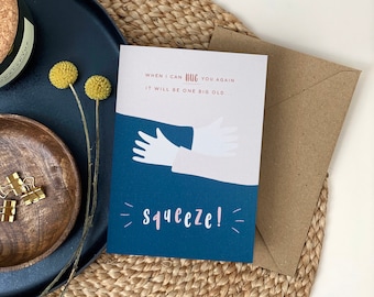 May include: A blue card with a pink and white illustration of two hands hugging. The text on the card reads "WHEN I CAN HUG YOU AGAIN IT WILL BE ONE BIG OLD...Squeeze!"