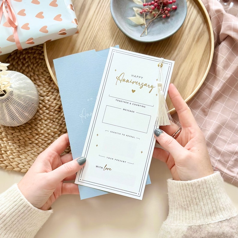 May include: White card with gold lettering that says "Happy Anniversary" and has a small gold heart. The card has a scratch-off area to reveal a message and a space for a gift. The card has a white tassel.