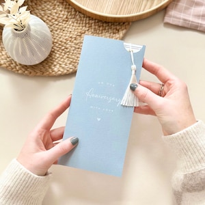 May include: A light blue card with the text "On our Anniversary with love" in white script. A small white heart is below the text. A white tassel hangs from the top of the card.