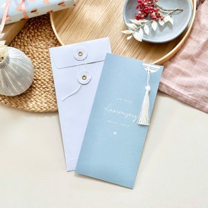 May include: A light blue card with white text that reads "On our Anniversary with love" and a small white heart. The card has a white tassel attached to it and is in a white envelope with gold buttons.