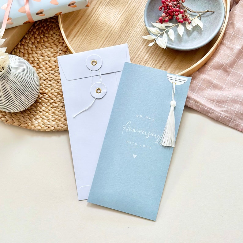 May include: A light blue card with white text that reads "On our Anniversary with love" and a small white heart. The card has a white tassel attached to it and is in a white envelope with gold buttons.