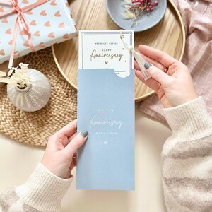 May include: A light blue card with the text "On our Anniversary with love" in white script. The card is folded in half and has a white tassel attached to the top. The card is being held by a person's hand.