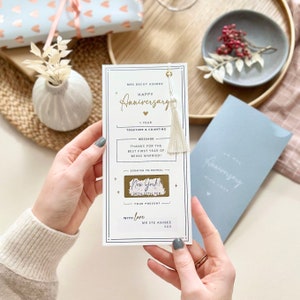 May include: A white card with a gold tassel and a gold scratch-off area. The card says "Happy Anniversary" and "1 year together & counting". The scratch-off area says "New York! 20th-27th Feb".