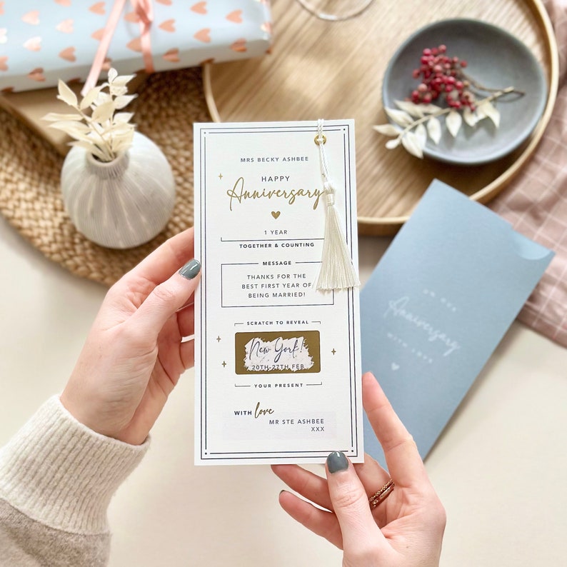 May include: A white card with a gold tassel and a gold scratch-off area. The card says "Happy Anniversary" and "1 year together & counting". The scratch-off area says "New York! 20th-27th Feb".