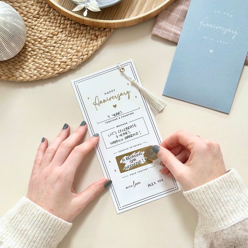 May include: A white card with gold lettering that says "Happy Anniversary" and "7 Years Together & Counting". The card has a scratch-off area that says "Let's Celebrate 7 Years Happily Married!" and reveals a message that says "A Relaxing Spa Weekend!" The card is decorated with gold stars and a small gold heart.