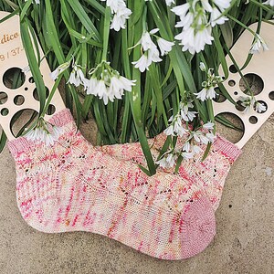 May include: A pair of hand-knitted socks in shades of pink, cream, and yellow, with a delicate lace pattern. The socks are displayed with a wooden sock form and surrounded by white flowers and green foliage.
