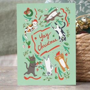 May include: A Christmas card with a pale green background, illustrated with cartoon cats in various poses. The cats wear Santa hats and are surrounded by festive decorations. The words "Yay Christmas" are written in red.