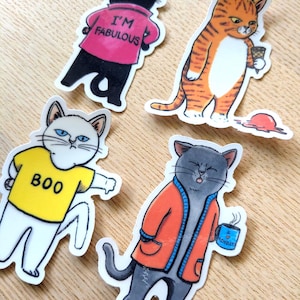 Op de afbeelding: Vier gestanste stickers met cartoonkatten. Een zwarte kat draagt een roze shirt met de tekst I'M FABULOUS. Een oranje cyperse kat houdt een gevallen ijshoorntje vast. Een witte kat draagt een geel shirt met de tekst BOO. Een grijze kat draagt een oranje vest en houdt een blauwe mok vast met de tekst I HATE MONDAYS.
