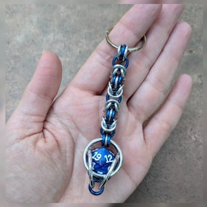 Puede incluir: Un llavero azul y plateado con un dije de dado azul y blanco. El llavero está hecho de un diseño de cadena de eslabones.