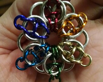 Puede incluir: Un colgante de flor de malla metálica de colores con base plateada y anillos en rojo, naranja, amarillo, verde, azul y morado.