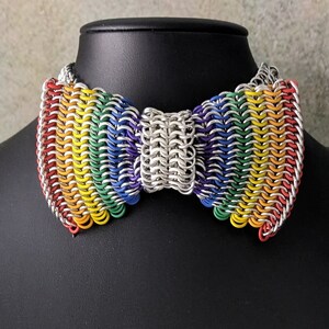 May include: A rainbow-coloured chainmail bow tie on a black display stand. The bow tie is made of small metal rings and is in the shape of a bow tie. The colours of the bow tie are red, orange, yellow, green, blue, indigo, and violet.