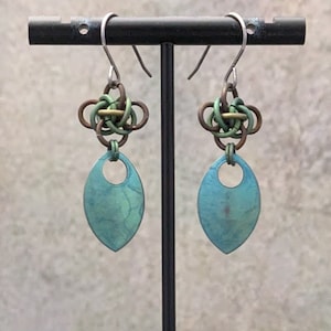 Könnte beinhalten: Ein Paar türkisblaue und grüne, blattartige Ohrringe mit einem geknüpften Metalldesign an der Oberseite. Die Ohrringe hängen an einem schwarzen Ohrringständer.