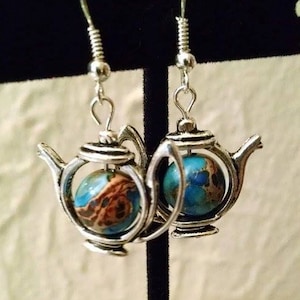 May include: Silver-toned teapot earrings. Each earring features a teapot-shaped frame encasing a round, blue and brown stone. The earrings hang from silver-colored ear wires, creating a unique and whimsical accessory.