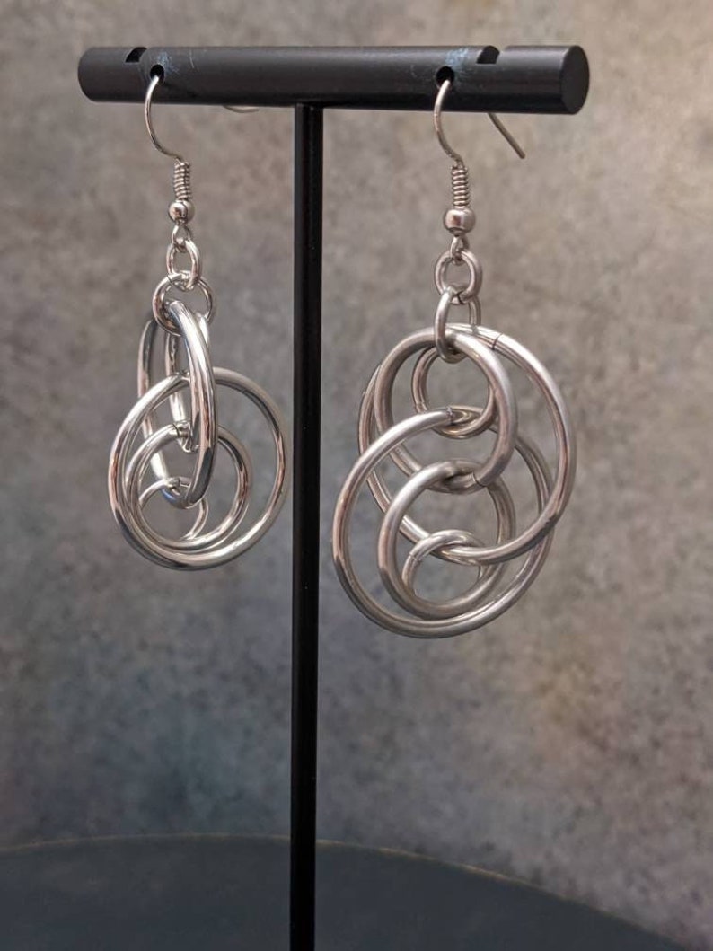 May include: Silver hoop earrings with multiple interlocking circles. The earrings are hanging from a black stand.