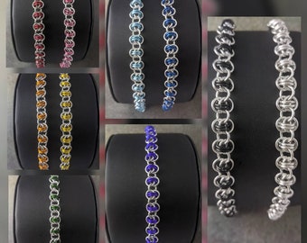 May include: A set of eight bracelets made of silver and colored metal rings. The bracelets are displayed on black stands. The colors of the bracelets are red, pink, yellow, orange, blue, light blue, green, and purple.