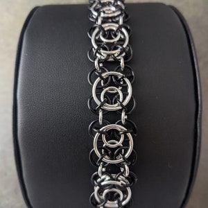 Puede incluir: Un brazalete de malla de cadena negro y plateado. El brazalete está hecho de pequeños anillos de metal que se entrelazan para crear un patrón de malla de cadena.