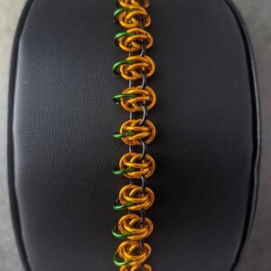 May include: A black, green, and orange chainmail bracelet. The bracelet is made of small metal rings that are linked together.