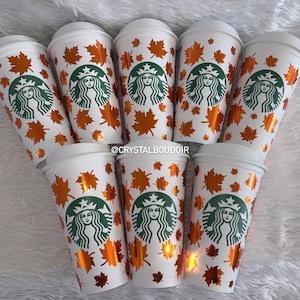 May include: Seven white reusable plastic cups with lids, each featuring a green Starbucks siren logo and a pattern of metallic orange maple leaves. The text CRYSTALBOUDOIR is printed in white across the centre of the image over the cups.