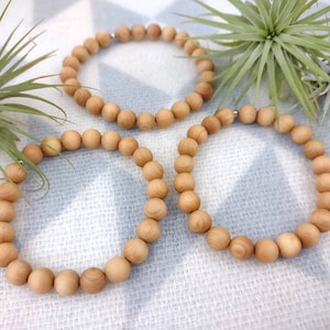 May include: Three wooden bead bracelets in various sizes. The bracelets are made of natural wood beads and are arranged on a white fabric background.