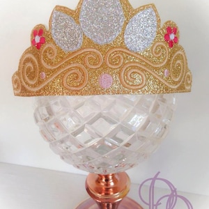 May include: A gold glitter princess crown with white and silver glitter accents. The crown has a pink and red flower on each side.