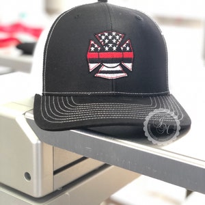 May include: Black and white trucker hat with a red line across the front. The hat features an embroidered American flag with a firefighter's cross.